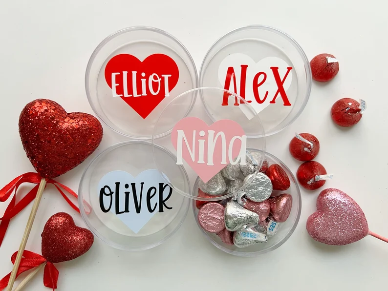Four clear plastic tubs with personalized heart decals (Nina, Elliot, Alex, Oliver) filled with Hershey's Kisses, serving as personalized Valentine's Day Party Favors. Etsy
