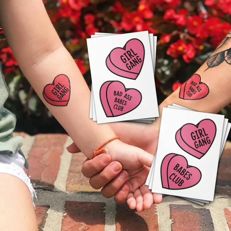 Heart-shaped temporary tattoos reading "GIRL GANG" and "BAD ASS BABES CLUB" shown on arms as playful Galentine's Day Party Favors.