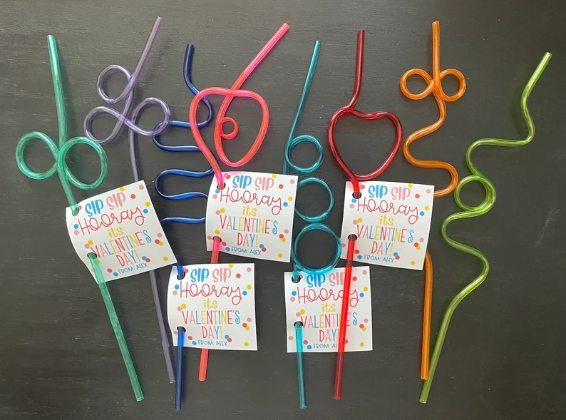 A colorful collection of twisted, heart-shaped silly straws, each attached with a small card reading "SIP SIP HOORAY, it's VALENTINE'S DAY!" as fun party favors.