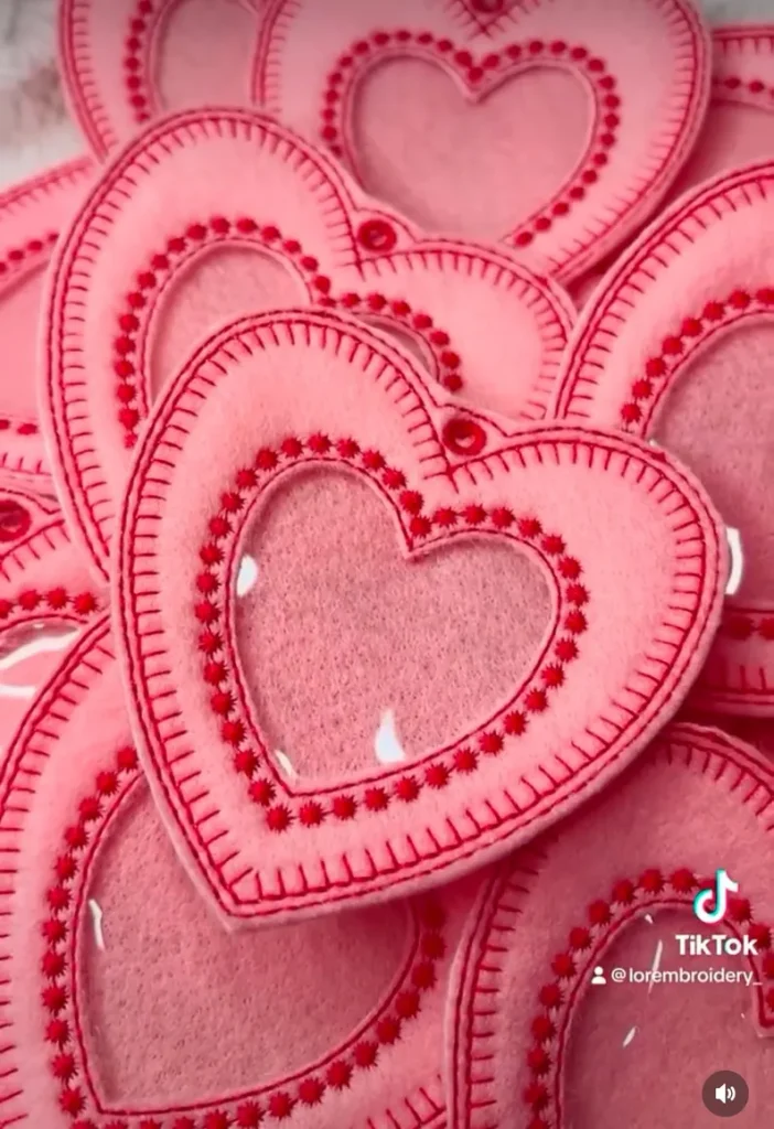 A pile of pink, heart-shaped felt pouches with red embroidered stitching and a clear vinyl window, unique Galentine's Day Party Favors.