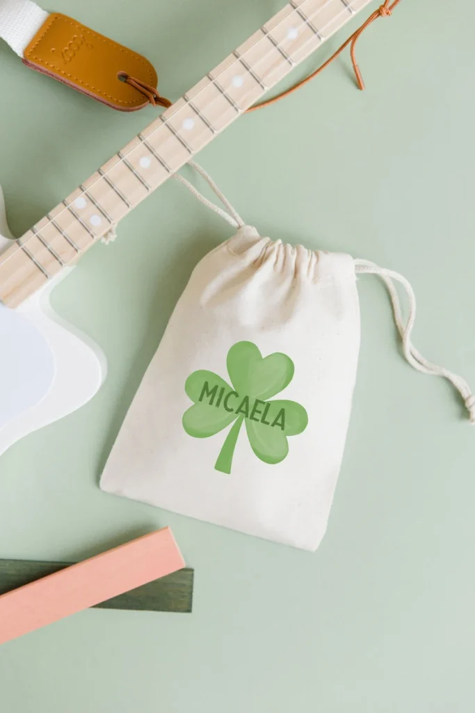 A small, cream-colored cotton drawstring bag personalized with a green shamrock and name, perfect for holding small and sustainable St. Patrick's Day favors.