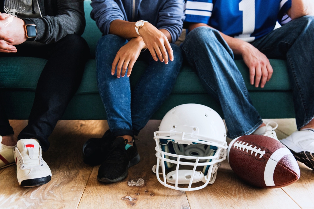 legs of football fans sitting on the couch with a helmet and american football at their feet