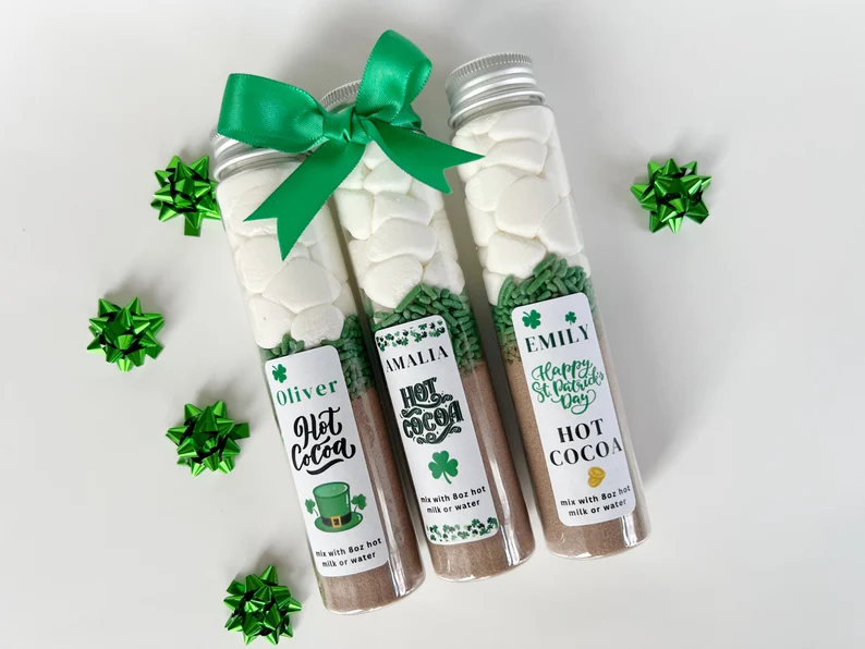 Personalized hot cocoa mix tubes layered with green sprinkles and marshmallows, topped with green ribbons as festive St. Patrick's Day favors.