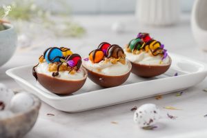 Three chocolate shells filled with creamy Easter egg cheesecakes, topped with colorful candy pieces and chocolate drizzle on a white tray.