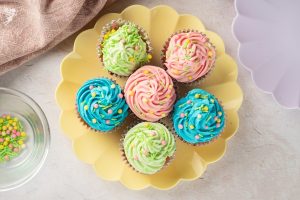 An overhead view of six Easter Pastel Vanilla Cupcakes with pink, blue, and green frosting arranged on a yellow scalloped plate.