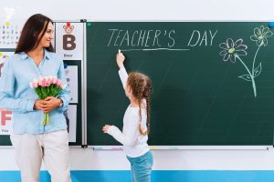 female teacher holding flowers with little girl writing teachers day on a blackboard with chalk