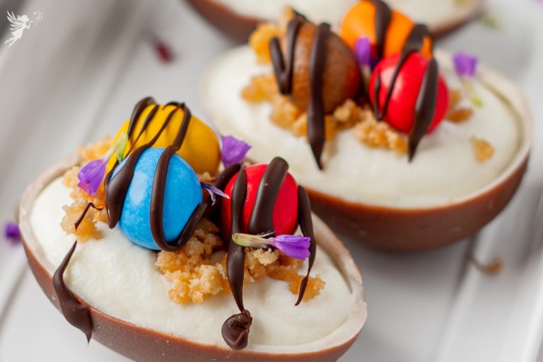 A close-up view of Easter egg cheesecakes featuring velvety filling, crunchy biscuit crumbs, vibrant candy eggs, and purple edible petals.