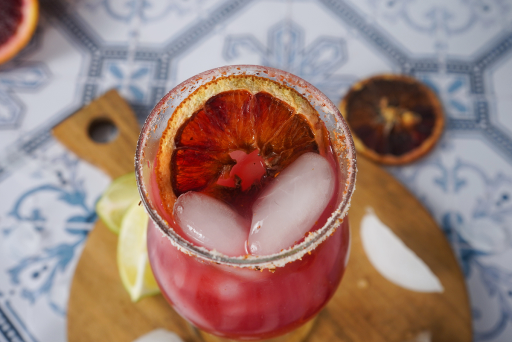 A chilled Blood Orange Margarita is served over ice in a salted glass, garnished with a beautiful dried citrus wheel.