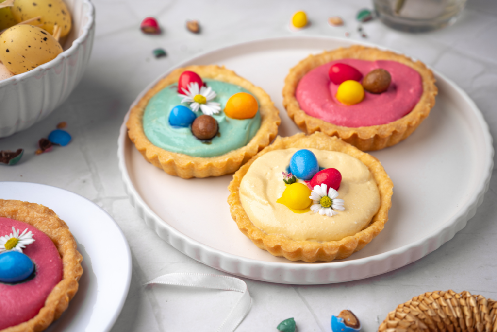 A group of mini spring cheesecake tarts with blue, yellow, and pink fillings, decorated with chocolate eggs for a kid-friendly Easter treat.