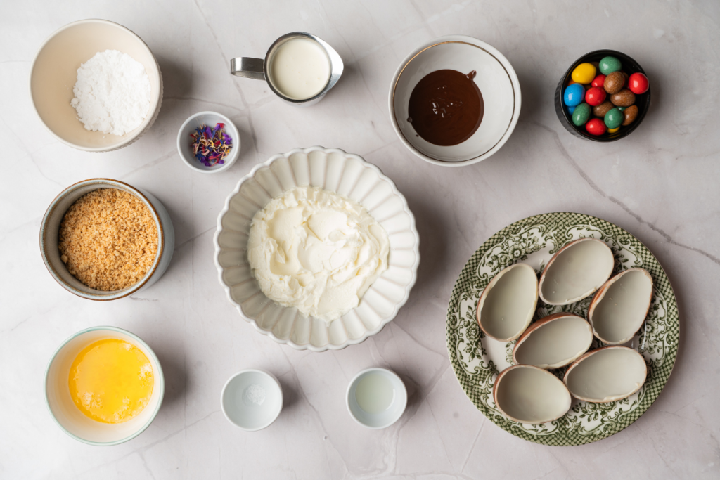 Flat lay of ingredients including cream cheese, biscuit crumbs, and chocolate shells to prepare homemade Easter egg cheesecakes.