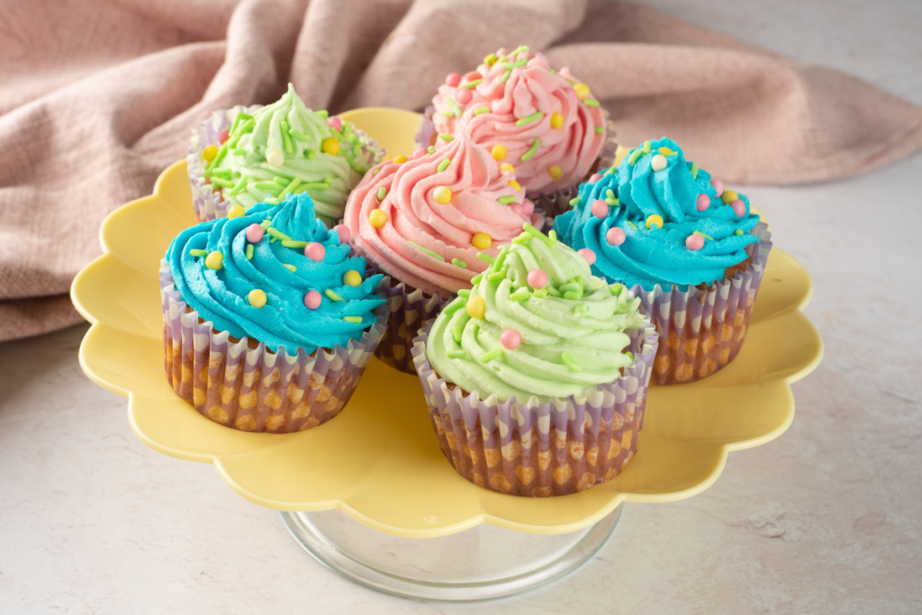 A yellow scalloped pedestal stand displaying finished Easter Pastel Vanilla Cupcakes topped with vibrant blue, pink, and green piped frosting.