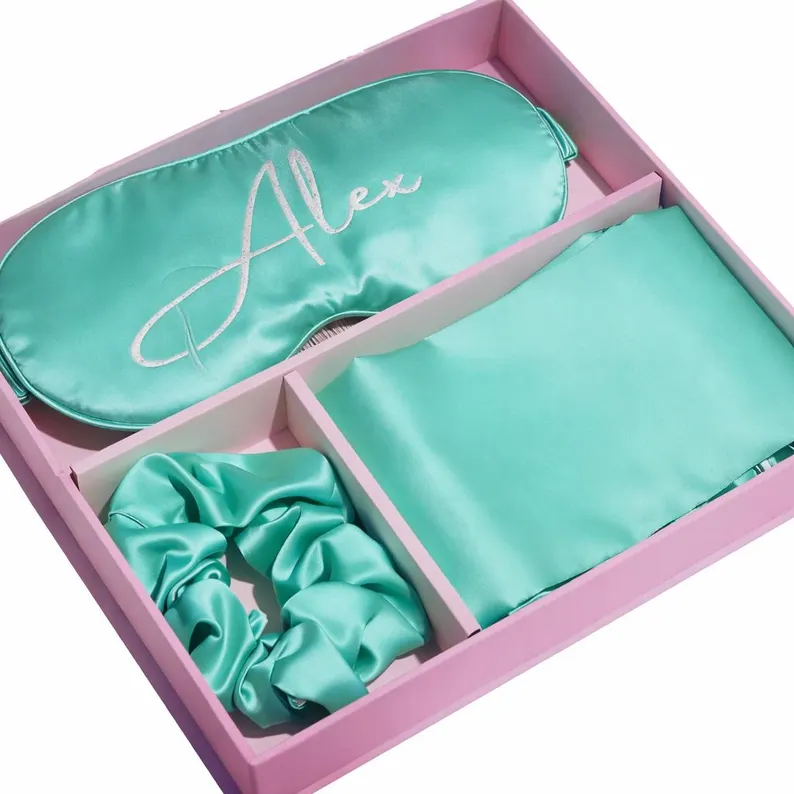 A pink divided gift box holding a teal satin pillowcase, scrunchie, and an embroidered eye mask for sleepover party favors.