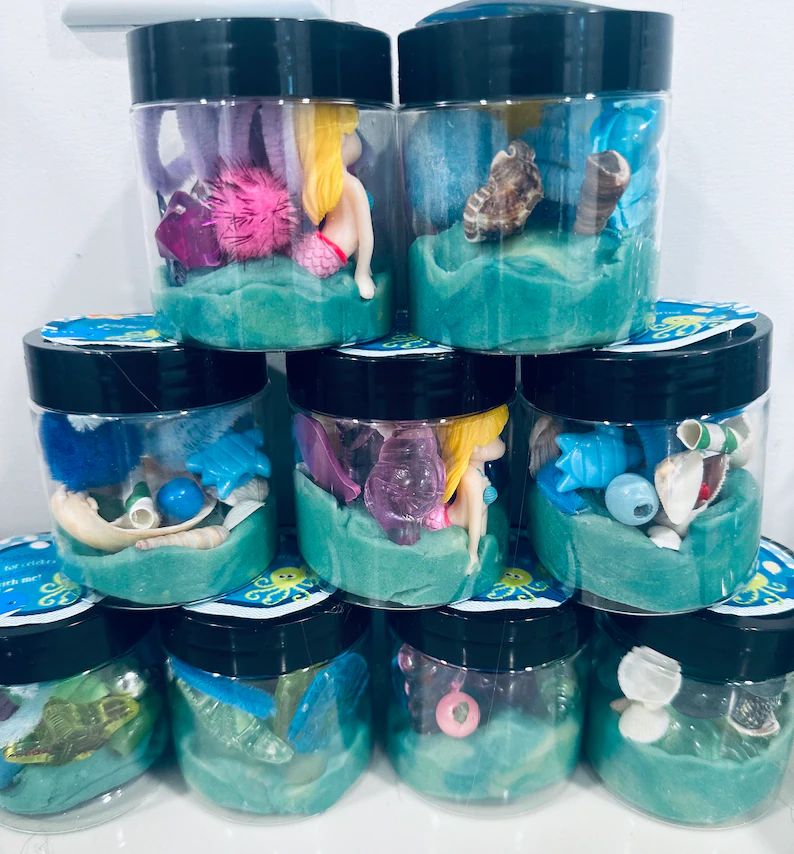 Under the sea play dough jars filled with turquoise dough and miniature ocean toys create interactive mermaid party favors for children's entertainment.