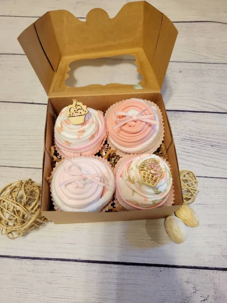 Four pink and white rolled baby bodysuit cupcakes with ribbon accents arranged in a kraft gift box, an adorable and creative baby shower gift presentation for newborn girls.
