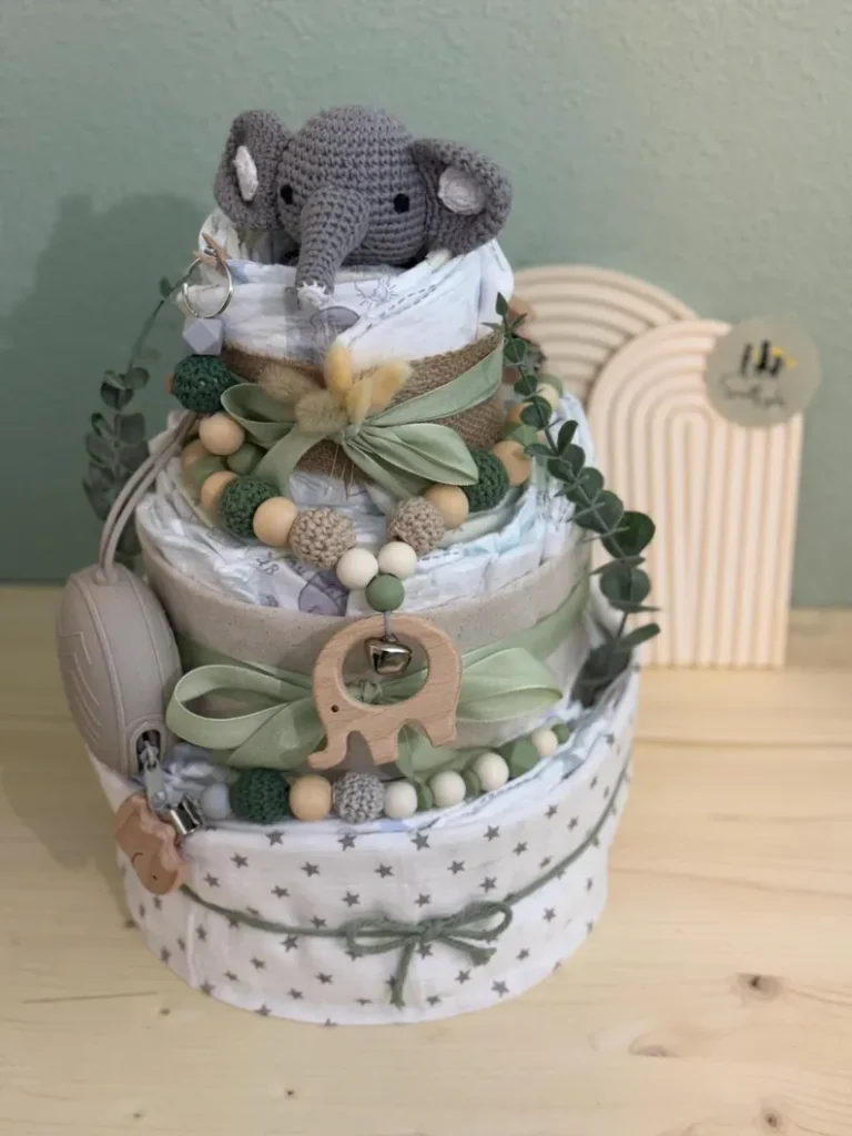 Three-tiered elephant-themed diaper cake topped with a crocheted elephant, decorated with wooden beads, green ribbons, and eucalyptus, a creative and practical baby shower gift.