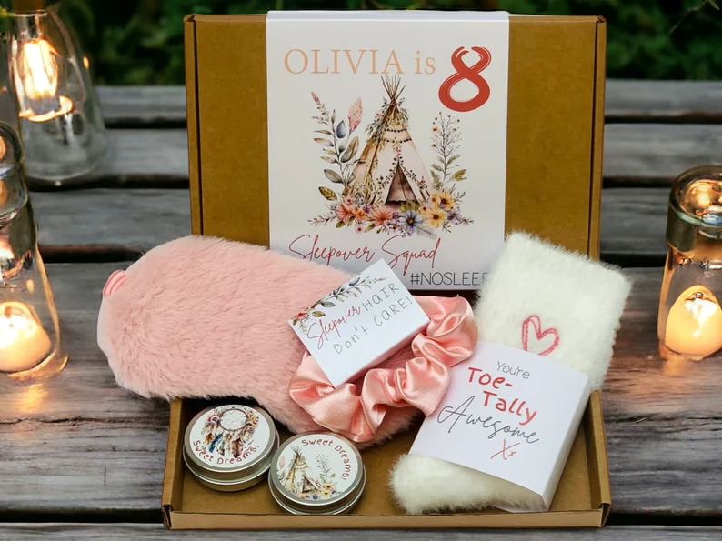 A cardboard gift box containing a pink fuzzy eye mask, white socks, and a satin scrunchie, perfect for sleepover party favors.