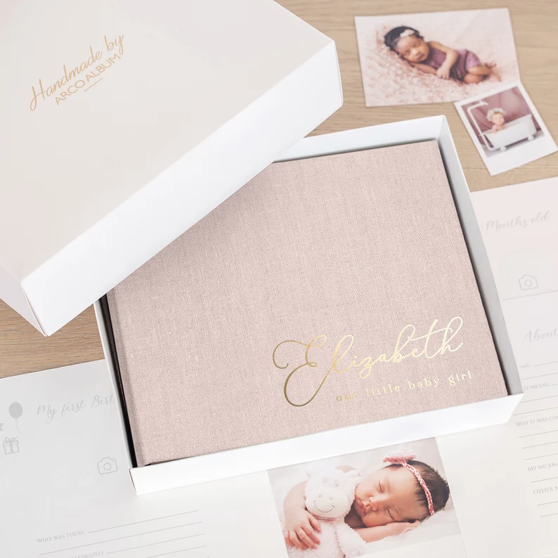 Personalized blush linen first year baby memory book with gold foil name Elizabeth displayed in a white gift box, an elegant keepsake baby shower gift for girls.