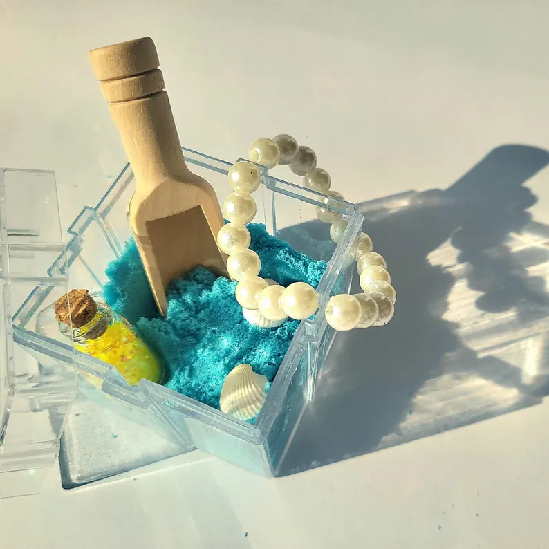 Clear kinetic sand box filled with turquoise sand, wooden shovel, pearl bracelet, and shells creates interactive sensory mermaid party favors.