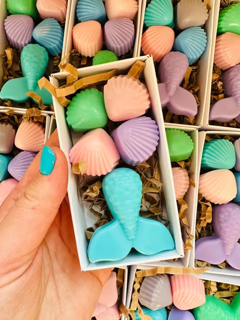 Handmade mermaid soaps shaped like shells and tails in pastel colors packaged in white boxes create delightful mermaid party favors for guests.
