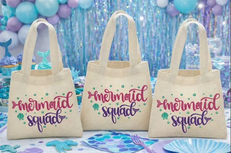 Canvas mermaid squad tote bags with pink and purple lettering and shell accents make practical and cute mermaid party favors for children.