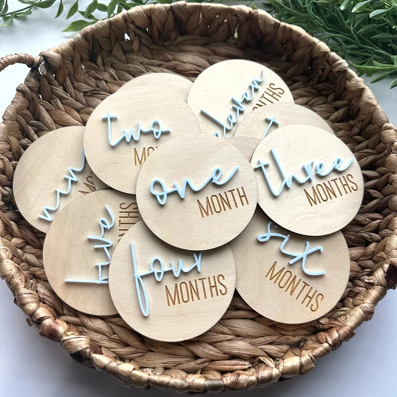 Wooden monthly milestone disc set with blue script lettering displayed in a wicker basket, meaningful baby shower gifts for capturing a newborn's first year.