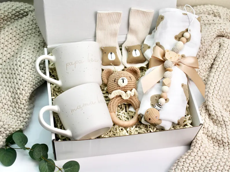 White gift box containing Mama Bear and Papa Bear mugs, bear booties, crochet rattle, muslin blanket, and wooden pacifier clip, charming baby shower gifts for new parents.