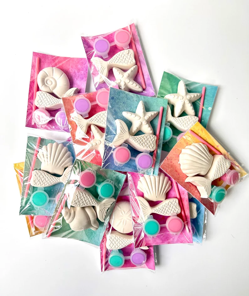 Ocean-themed paint kits with mermaid tails, seashells, and starfish figures in vibrant tie-dye bags make creative mermaid party favors for kids. Etsy
