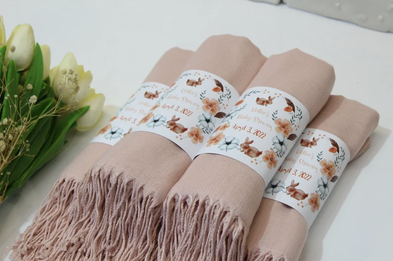 Four blush pink pashmina shawls rolled and wrapped with personalized floral bunny-themed belly bands for Lottie's Baby Shower, practical and beautiful baby shower party favors. Etsy