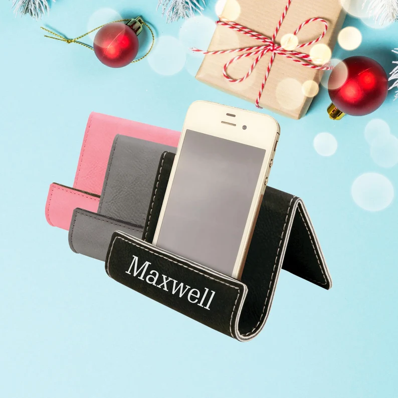 Custom engraved leather phone holder in multiple colors, thoughtful tween teen boy party favors displayed with festive Christmas decorations. Etsy