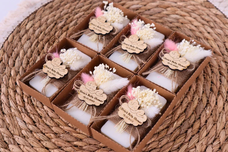 Six white scented soaps decorated with dried flowers, pink pampas grass, and personalized wooden tags reading Olivia's Baby Shower, rustic boho baby shower party favors arranged in a kraft box. Etsy