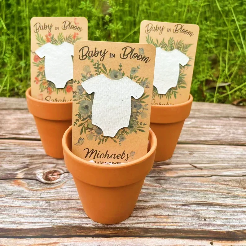 Three personalized "Baby in Bloom" seed paper onesie cards displayed in terracotta pots, eco-friendly and plantable baby shower party favors for nature-themed celebrations. Etsy