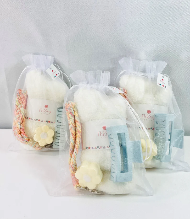 Three white organza bags containing fuzzy socks, a blue hair claw clip, and a flower bath bomb as sleepover party favors.