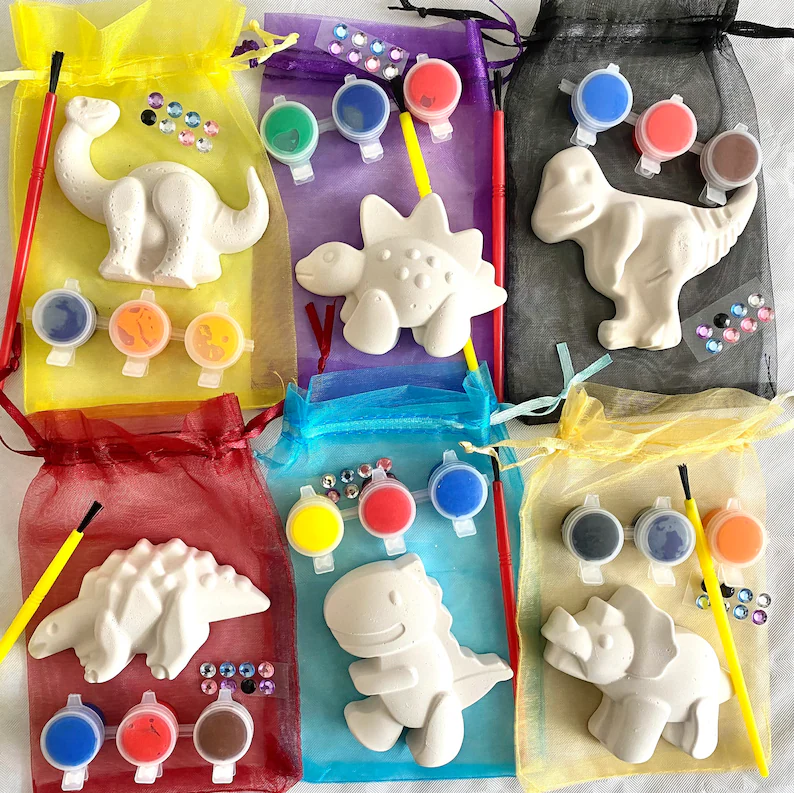 Dinosaur craft kit dino party favors in colorful organza bags, each containing unpainted ceramic dinosaur figurines, paint pots, brushes, and decorative gems for creative activities. Etsy