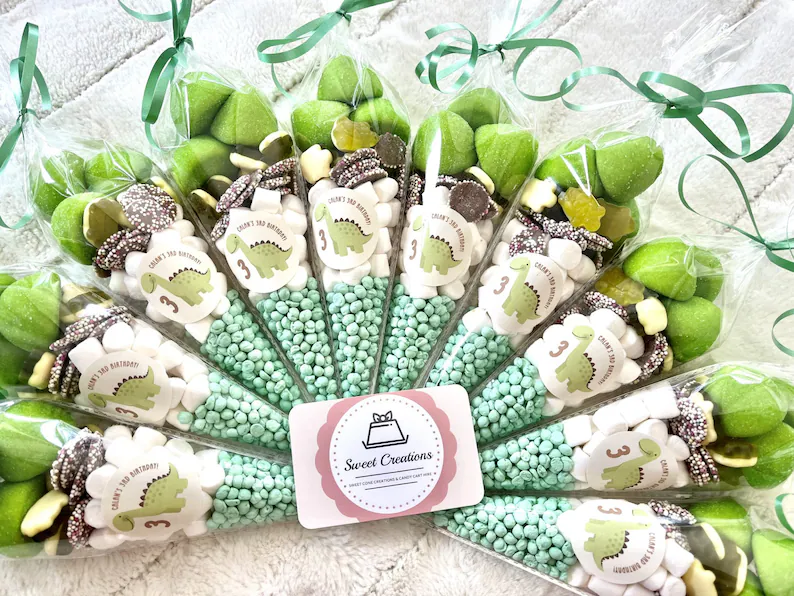 Dinosaur sweetie cones dino party favors arranged in fan display, filled with mint green candy pearls and marshmallows, decorated with personalized dinosaur labels and green ribbons. Etsy