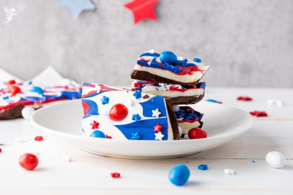 Stack of Red White & Blue Chocolate Bark pieces decorated with swirled patriotic colors and candy toppings on white plate with star decorations.