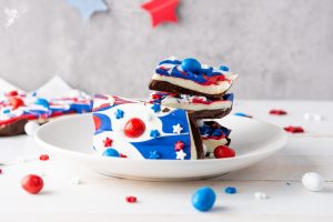 Stack of Red White & Blue Chocolate Bark pieces decorated with swirled patriotic colors and candy toppings on white plate with star decorations.