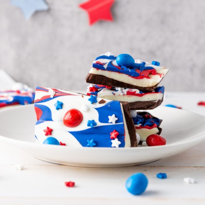 Stack of Red White & Blue Chocolate Bark pieces decorated with swirled patriotic colors and candy toppings on white plate with star decorations.