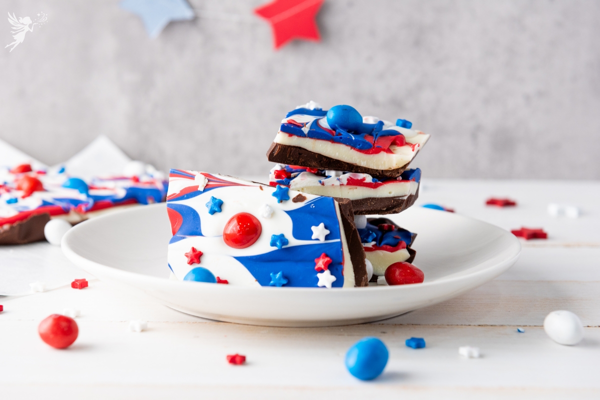 Stack of Red White & Blue Chocolate Bark pieces decorated with swirled patriotic colors and candy toppings on white plate with star decorations.