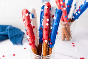Patriotic Red White Blue Firecracker Pretzel Rods dipped in colored chocolate with star sprinkles displayed in glass jar on white marble surface.