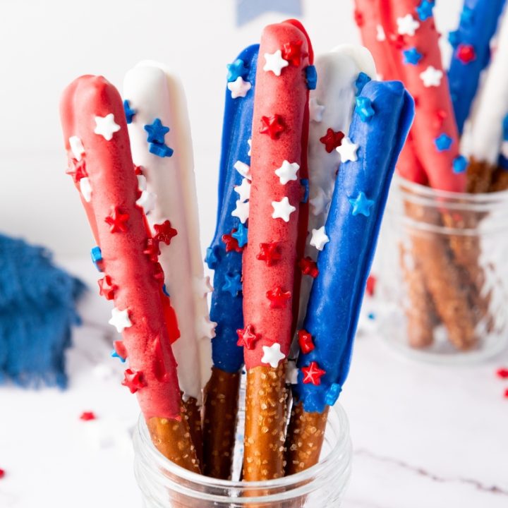 Patriotic Red White Blue Firecracker Pretzel Rods dipped in colored chocolate with star sprinkles displayed in glass jar on white marble surface.