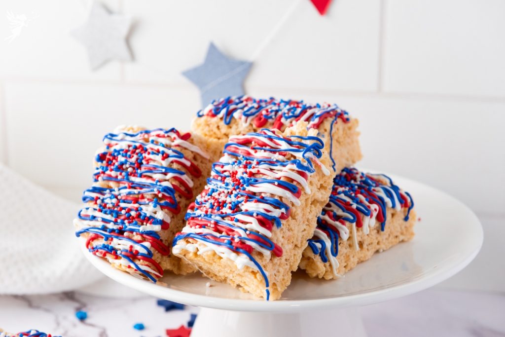 Stack of Patriotic Rice Krispie Treats decorated with red, white, and blue candy drizzle and sprinkles on a white cake stand with star decorations.
