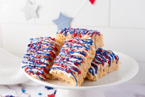 Stack of Patriotic Rice Krispie Treats decorated with red, white, and blue candy drizzle and sprinkles on a white cake stand with star decorations.