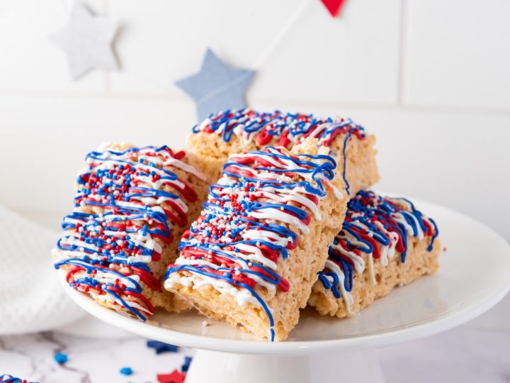 Stack of Patriotic Rice Krispie Treats decorated with red, white, and blue candy drizzle and sprinkles on a white cake stand with star decorations.