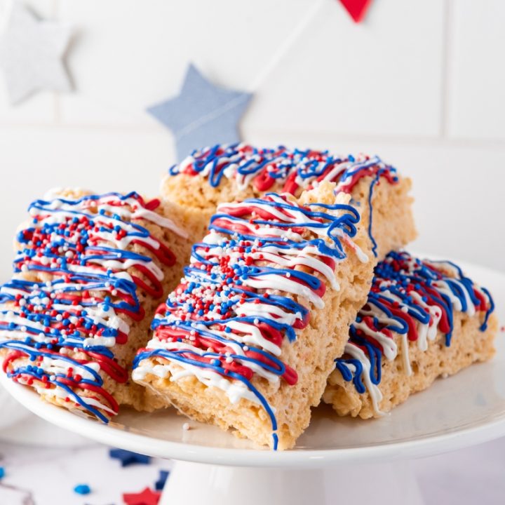 Stack of Patriotic Rice Krispie Treats decorated with red, white, and blue candy drizzle and sprinkles on a white cake stand with star decorations.