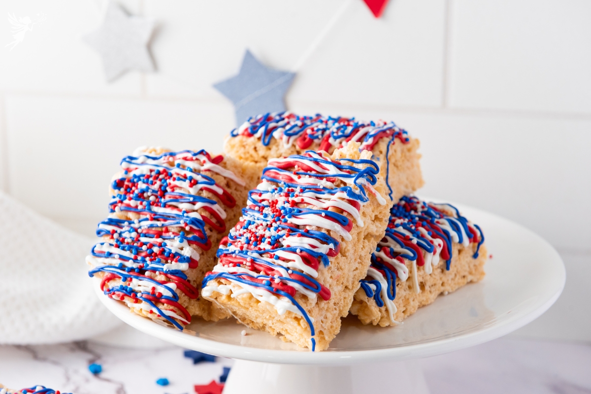 Stack of Patriotic Rice Krispie Treats decorated with red, white, and blue candy drizzle and sprinkles on a white cake stand with star decorations.