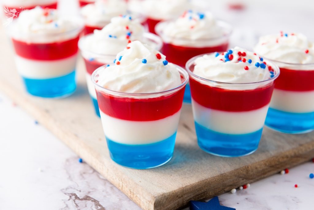 Non Alcoholic Red White & Blue Jello Shots layered in clear cups with whipped cream and patriotic sprinkles on wooden board—perfect 4th of July and Memorial Day dessert.