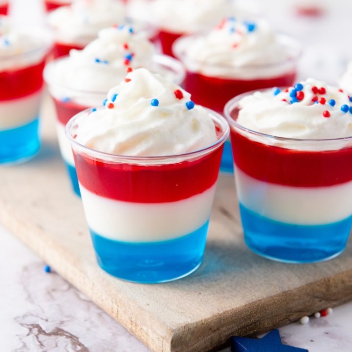 Non Alcoholic Red White & Blue Jello Shots layered in clear cups with whipped cream and patriotic sprinkles on wooden board—perfect 4th of July and Memorial Day dessert.