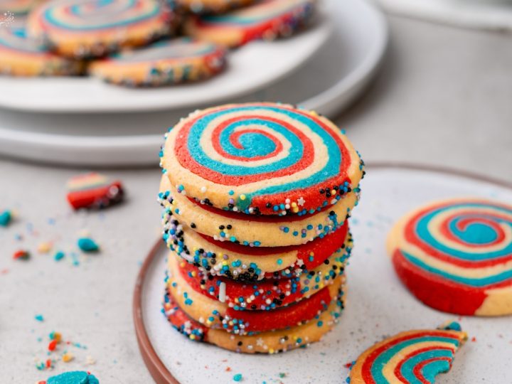 Stack of red white and blue patriotic pinwheel cookies with spiral swirl pattern and colorful sprinkle edges on plate.