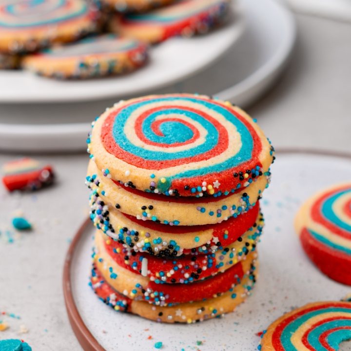 Stack of red white and blue patriotic pinwheel cookies with spiral swirl pattern and colorful sprinkle edges on plate.