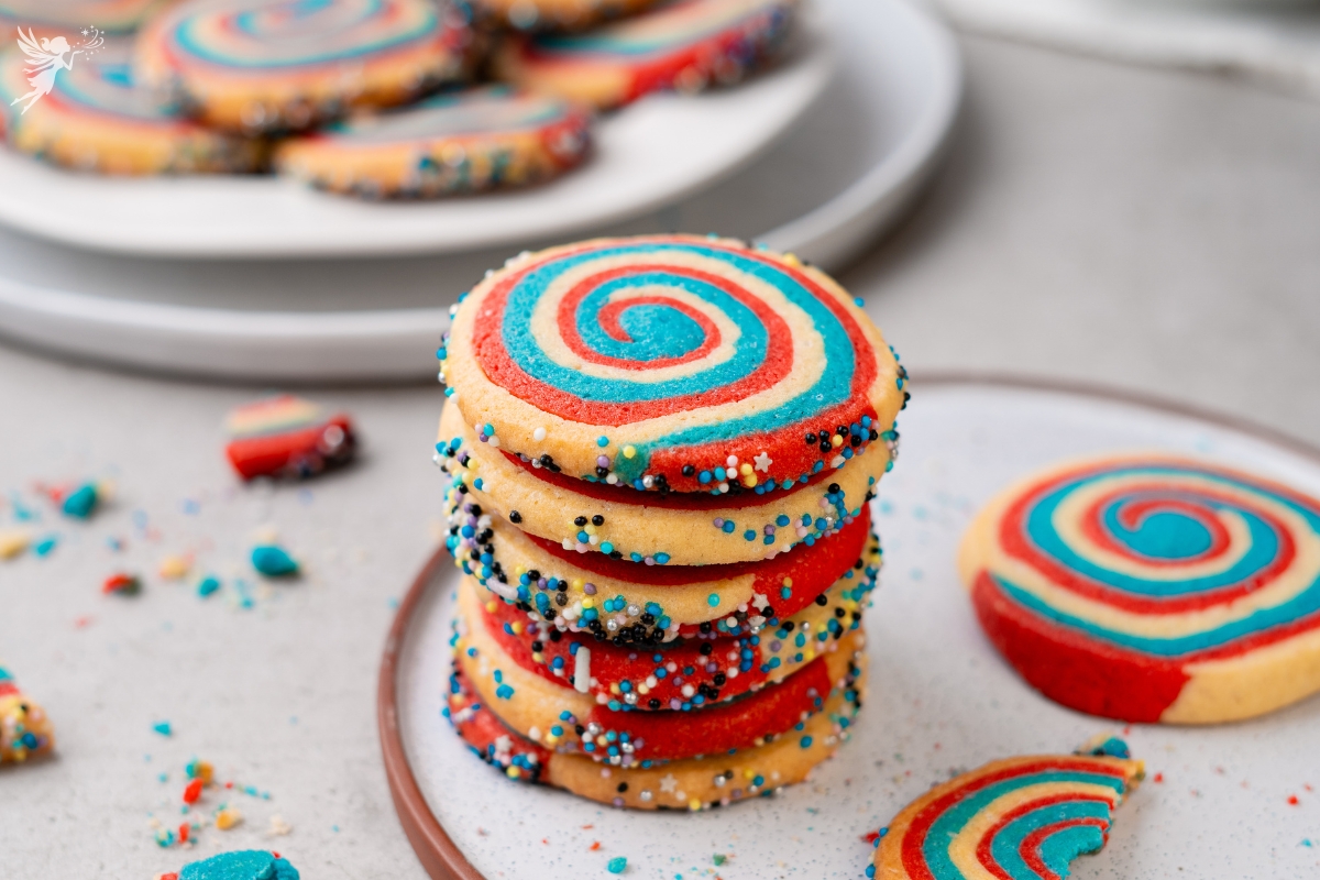 Stack of red white and blue patriotic pinwheel cookies with spiral swirl pattern and colorful sprinkle edges on plate.
